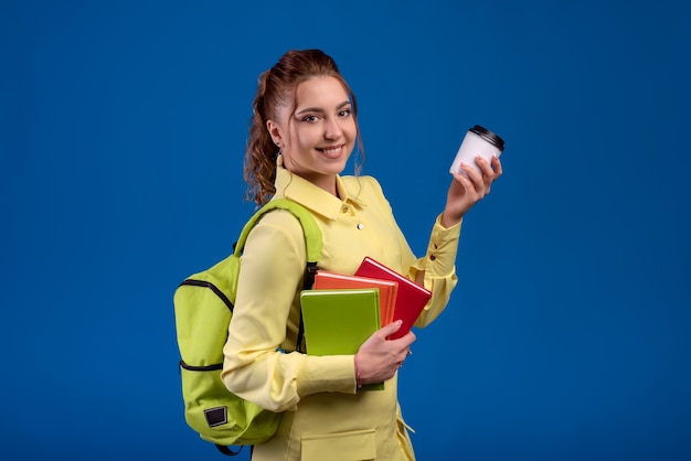 Mulher casual segurando um copo de cappuccino descartável e segurando uma mochila isolada na parede azul