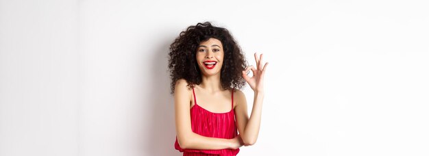 Mulher caucasiana alegre com cabelo encaracolado e maquiagem usando vestido vermelho elegante mostrando sinal ok e sm