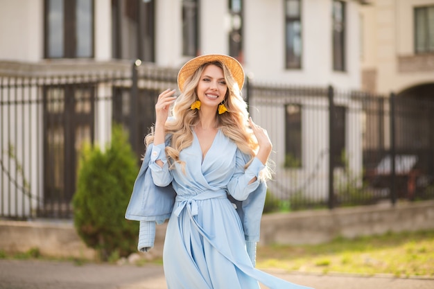 Mulher caucasiana alegre com longos cabelos loiros, olhos azuis e um sorriso perfeito, em um vestido longo azul com um chapéu, sai perto dos edifícios