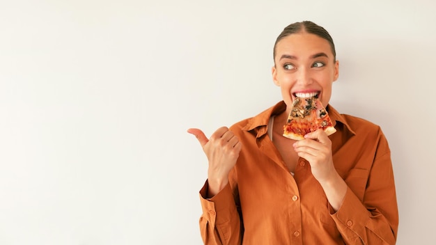 Foto mulher caucasiana alegre mordendo uma fatia de pizza saborosa e apontando de lado para o espaço livre na parede branca