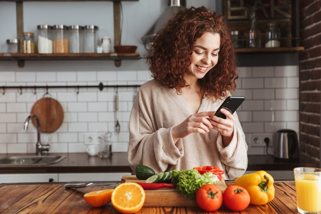 Mulher caucasiana alegre segurando um smartphone enquanto cozinha salada com legumes frescos no interior da cozinha em casa