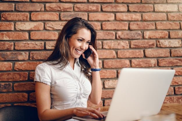 Mulher caucasiana alegre sorridente usando o laptop e telefone inteligente enquanto está sentado no café. Conceito de multitarefa.