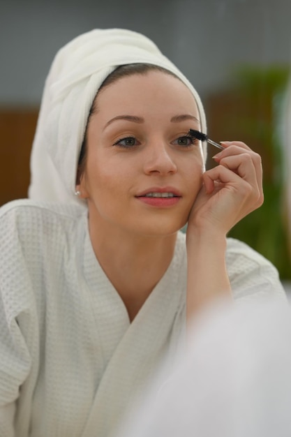 Mulher caucasiana atraente em roupão aplicando rímel nos cílios Conceito de beleza e cosméticos