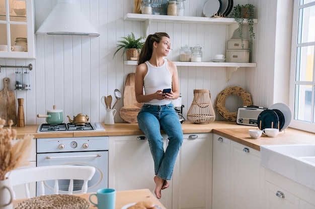 Mulher caucasiana calma com cabelo solto vestido com camiseta branca e jeans azul sentado na mesa da cozinha