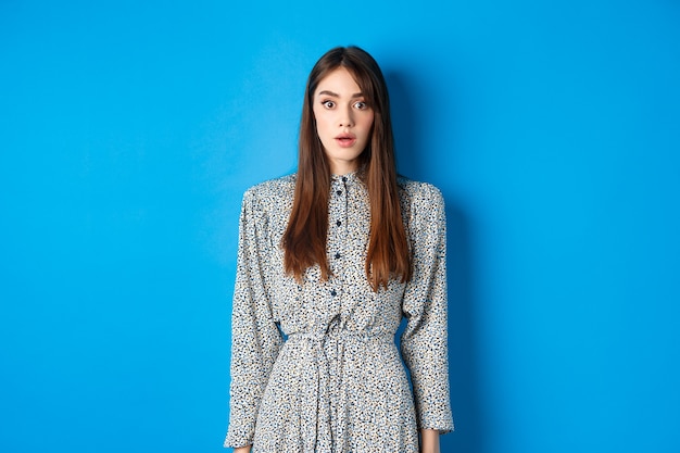 Mulher caucasiana chocada com cabelo comprido, usando vestido, ofegando e em pé no estúpido contra o azul, olha com descrença.