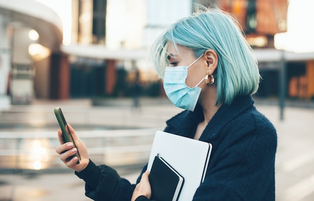 Mulher caucasiana com cabelo azul está conversando com alguém enquanto usa uma máscara protetora e segura um computador do lado de fora