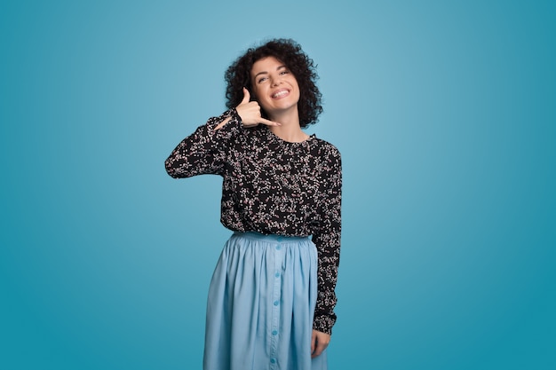 Mulher caucasiana com cabelo encaracolado, usando um vestido azul e posando em uma parede, gesticulando com o sinal de chamada sorrindo