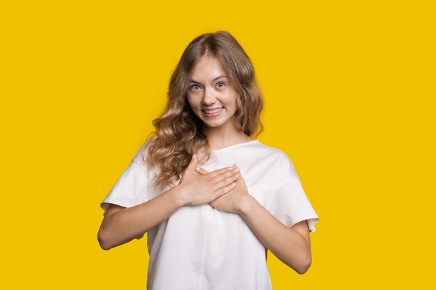 Mulher caucasiana com cabelo loiro e camiseta branca sorrindo em uma parede amarela tocando seu coração com as palmas das mãos