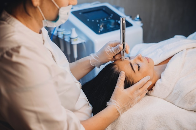 Mulher caucasiana com cabelo preto deitada na cama e submetida a um procedimento de cuidados da pele facial com aparelhos no salão spa