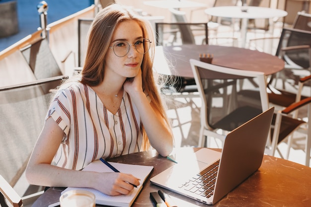 Mulher caucasiana com cabelo ruivo e sardas fazendo uma pausa para o café em uma cafeteria enquanto usa um laptop e faz algumas anotações