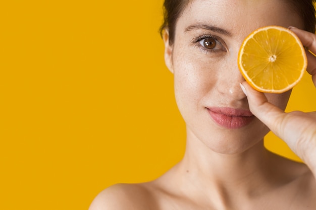 Mulher caucasiana com ombros nus posando com uma laranja cobrindo os olhos em uma parede amarela com espaço livre