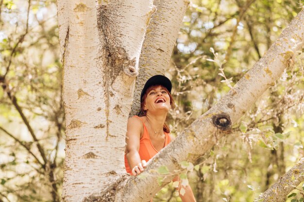 Foto mulher caucasiana com um boné sentado em uma árvore, rindo com uma expressão feliz no meio de uma floresta.