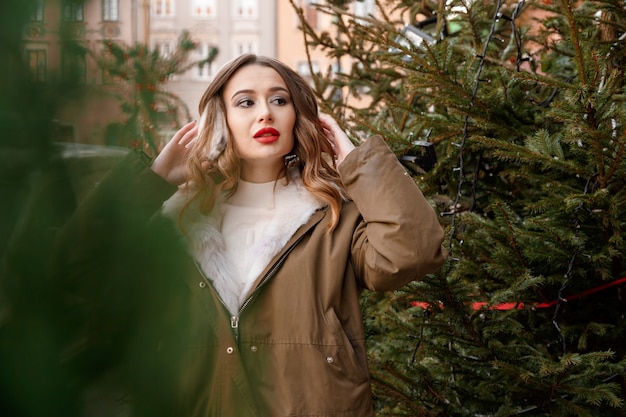 Mulher caucasiana com uma camisola de malha e fones de ouvido macios. Retrato ao ar livre de uma jovem linda feliz sorridente está vestindo um casaco de pele perto da árvore de Natal decorada. Polônia