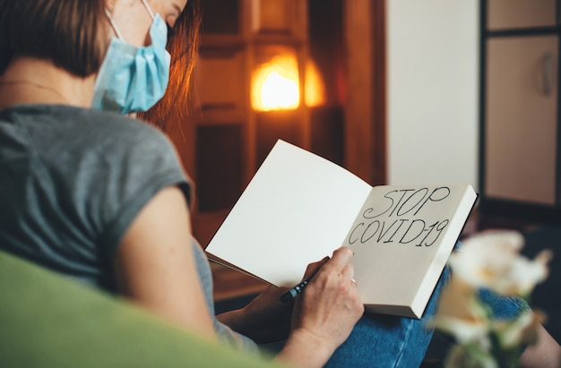 Foto mulher caucasiana com uma máscara especial está escrevendo algo em um livro enquanto está sentada no sofá