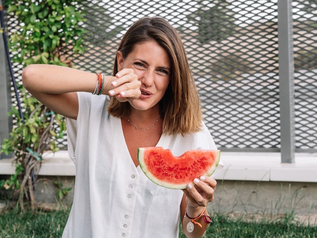 Mulher caucasiana, comendo fatias de melancia no jardim da casa dela e sentada no chão.