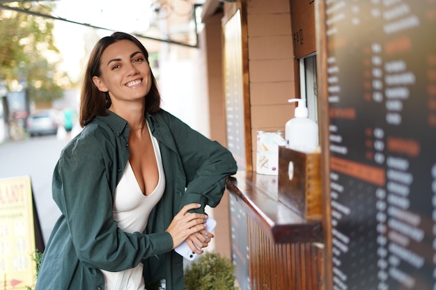 Foto mulher caucasiana comprando café na cafeteria, janela ao ar livre, hora do pôr do sol, sorrindo