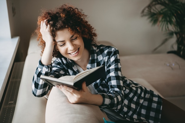 Mulher caucasiana de cabelos cacheados sorrindo no sofá enquanto lê um livro perto da janela