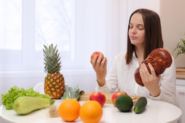 Mulher caucasiana de cabelos castanhos, pensativa e duvidosa, vestindo camisa branca, sentada à mesa, segurando uma maçã e uma torta, pensando no que comer, precisa manter a dieta, estilo de vida saudável