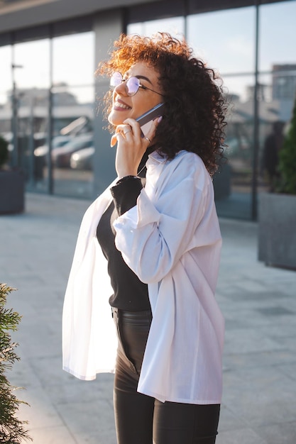 Mulher caucasiana de cabelos encaracolados falando no celular ao ar livre usando aparelhos modernos para comunicação