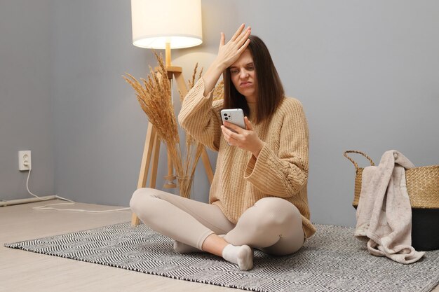 Foto mulher caucasiana de cabelos escuros infeliz vestindo roupas casuais usando telefone para trabalho remoto esquecida de uma tarefa importante fazendo gesto de palma facial sentada no chão no interior da casa