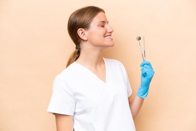 Mulher caucasiana de dentista segurando ferramentas isoladas em fundo bege rindo em posição lateral