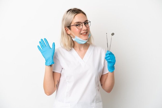 Mulher caucasiana de dentista segurando ferramentas isoladas no fundo branco e saudando com a mão com expressão feliz