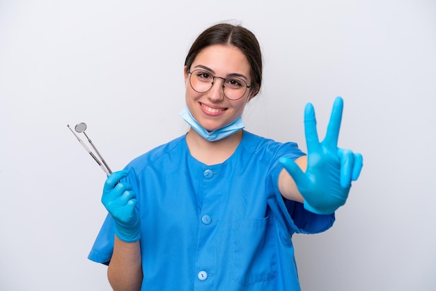 Mulher caucasiana de dentista segurando ferramentas isoladas no fundo branco feliz e contando três com os dedos