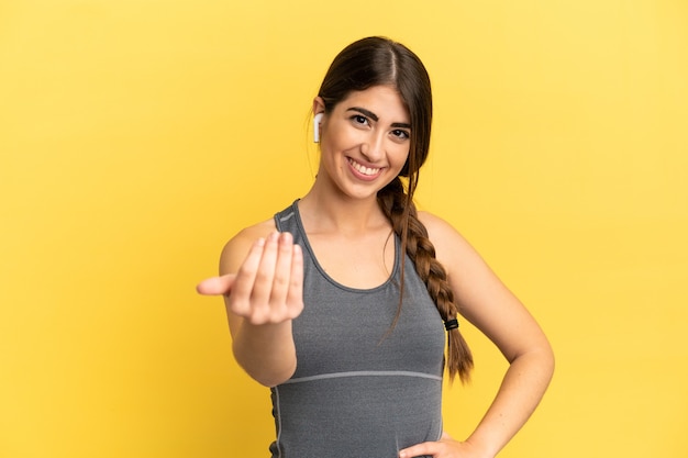 Mulher caucasiana de esporte isolada em um fundo amarelo, convidando para vir com a mão. Feliz que você veio