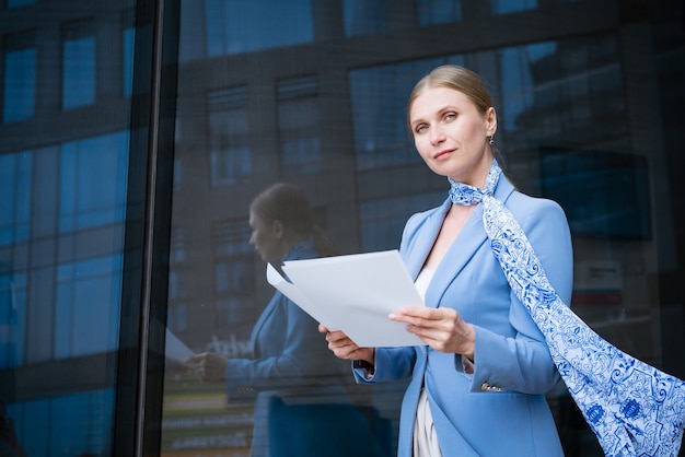 Mulher caucasiana de jaqueta azul e negócios bem-sucedidos segurando documentos perto de prédio de escritórios