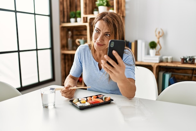 Mulher caucasiana de meia-idade comendo sushi usando smartphone em casa.