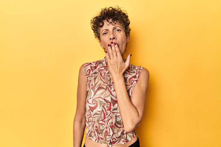 Foto mulher caucasiana de meia idade em amarelo vibrante bocejando mostrando um gesto cansado cobrindo a boca com a mão