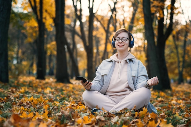 Mulher caucasiana de meia idade em fones de ouvido, ouvindo música, app de meditação no smartphone e meditando na posição de lótus no outono park. App de meditação, saúde mental, autocuidado, conceito de atenção plena.