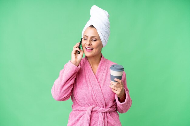 Mulher caucasiana de meia-idade em um roupão de banho sobre fundo isolado, mantendo uma conversa com o telefone móvel