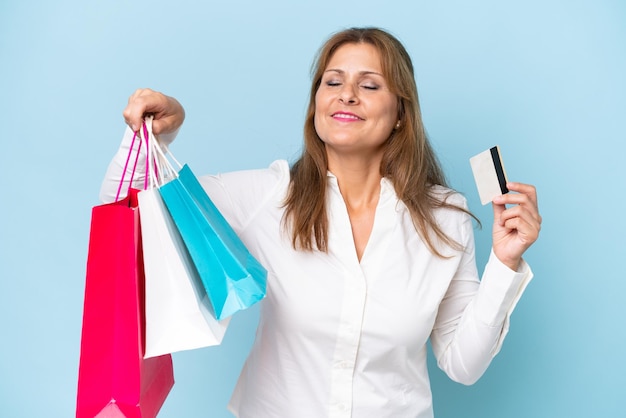 Mulher caucasiana de meia-idade isolada em fundo azul segurando sacolas de compras e um cartão de crédito