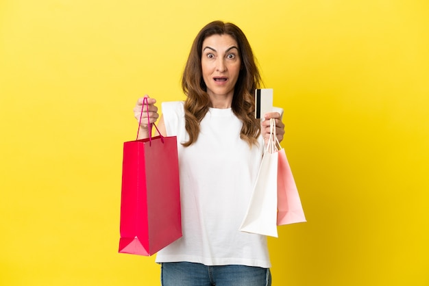 Mulher caucasiana de meia idade isolada em um fundo amarelo, segurando sacolas de compras e surpreso