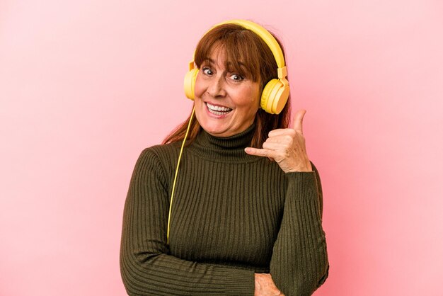 Foto mulher caucasiana de meia-idade, ouvindo música isolada no fundo rosa, mostrando um gesto de chamada de celular com os dedos.