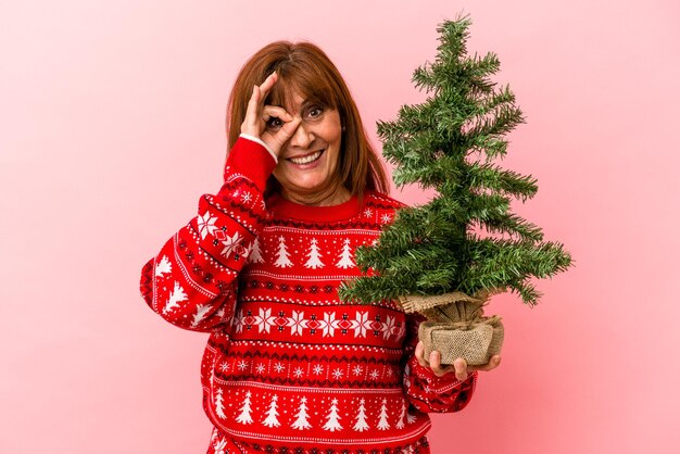 Mulher caucasiana de meia idade segurando a árvore de Natal isolada no fundo rosa, animado, mantendo o gesto ok no olho.