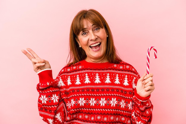 Mulher caucasiana de meia idade segurando a vara de natal isolada no fundo rosa alegre e despreocupada, mostrando um símbolo de paz com os dedos.