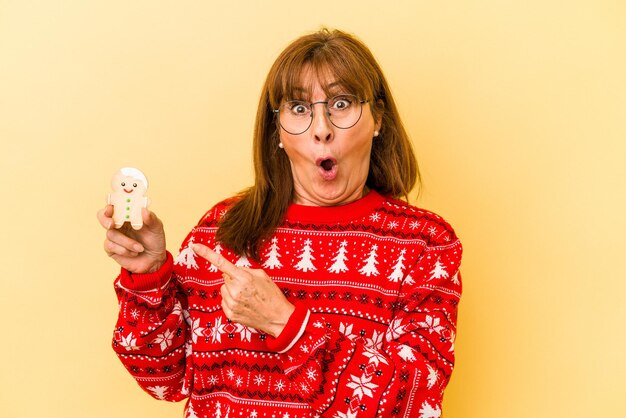 Mulher caucasiana de meia-idade segurando biscoitos de Natal isolados em um fundo amarelo apontando para o lado