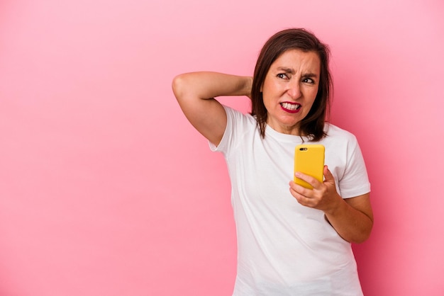 Mulher caucasiana de meia-idade segurando o celular isolado no fundo rosa tocando a parte de trás da cabeça, pensando e fazendo uma escolha.