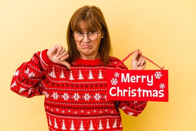 Mulher caucasiana de meia-idade segurando um cartaz de 'feliz natal' isolado em um fundo amarelo, mostrando um gesto de desagrado, polegar para baixo. conceito de desacordo.