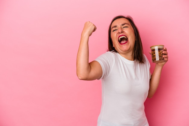 Foto mulher caucasiana de meia idade segurando um frasco de amêndoa isolado no fundo rosa, levantando o punho após uma vitória, o conceito de vencedor.