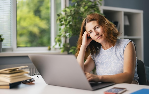 Mulher caucasiana de meia idade sonhando com fins de semana enquanto trabalhava no laptop moderno em casa Freelancer feminino usando óculos e roupas casuais