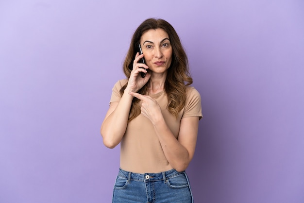 Mulher caucasiana de meia idade usando telefone celular isolado em um fundo roxo apontando para o lado para apresentar um produto