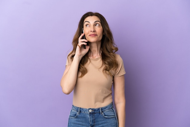 Mulher caucasiana de meia idade usando telefone celular isolado em um fundo roxo, olhando para o lado e sorrindo