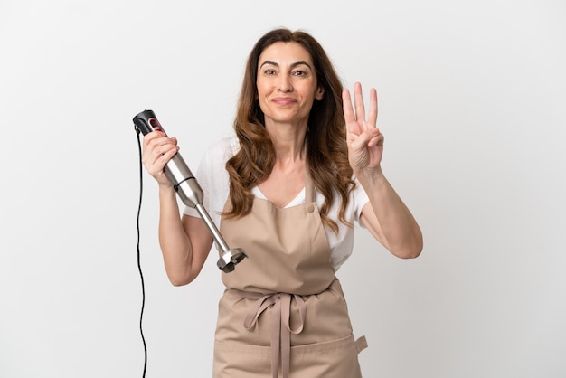 Mulher caucasiana de meia idade usando um liquidificador isolado no fundo branco feliz e contando três com os dedos