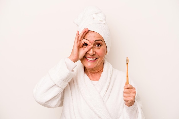 Mulher caucasiana de meia-idade vestindo um roupão de banho segurando a escova de dentes isolada no fundo branco animada mantendo o gesto okey no olho