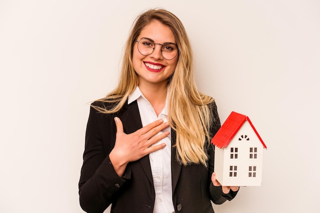 Foto mulher caucasiana de negócios jovem segurando uma casa de brinquedo isolada no fundo branco ri alto, mantendo a mão no peito