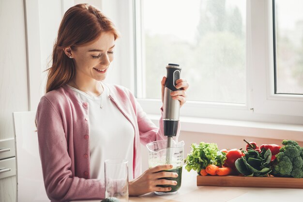 Mulher caucasiana de sardas ruivas espremendo suco de vegetais com um espremedor sorrindo na cozinha