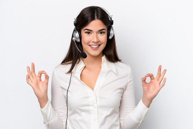 Mulher caucasiana de telemarketing trabalhando com um fone de ouvido isolado no fundo branco, mostrando um sinal de ok com os dedos